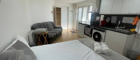A compact hotel room with a bed, a sofa, a small table, a washing machine, and a kitchenette.