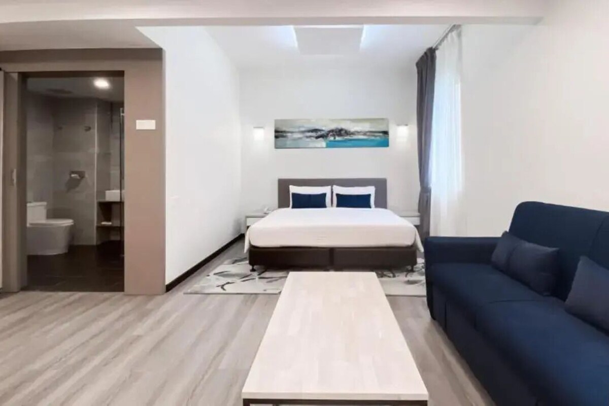 A modern hotel room with a bed, a sofa, a small table, and a shower area.