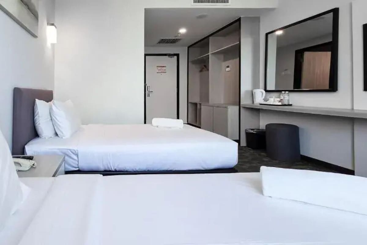 A hotel room with a bed, a bedside table with a phone, a mirror, a wardrobe, and a trash bin.