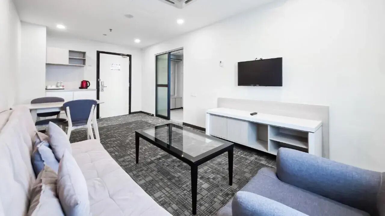 Ein modernes Hotelzimmer mit Glastisch, einer Couch, einem an der Wand montierten Fernseher und einer Küchenzeile.