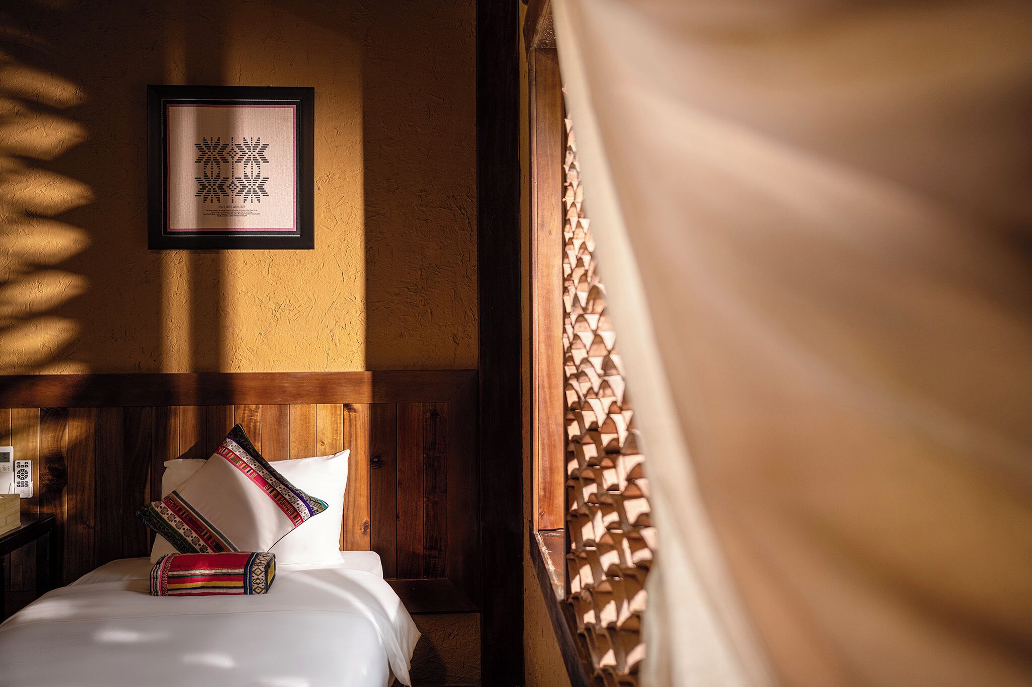 A hotel room with a bed, pillows, a framed picture on the wall, and a window with a curtain.
