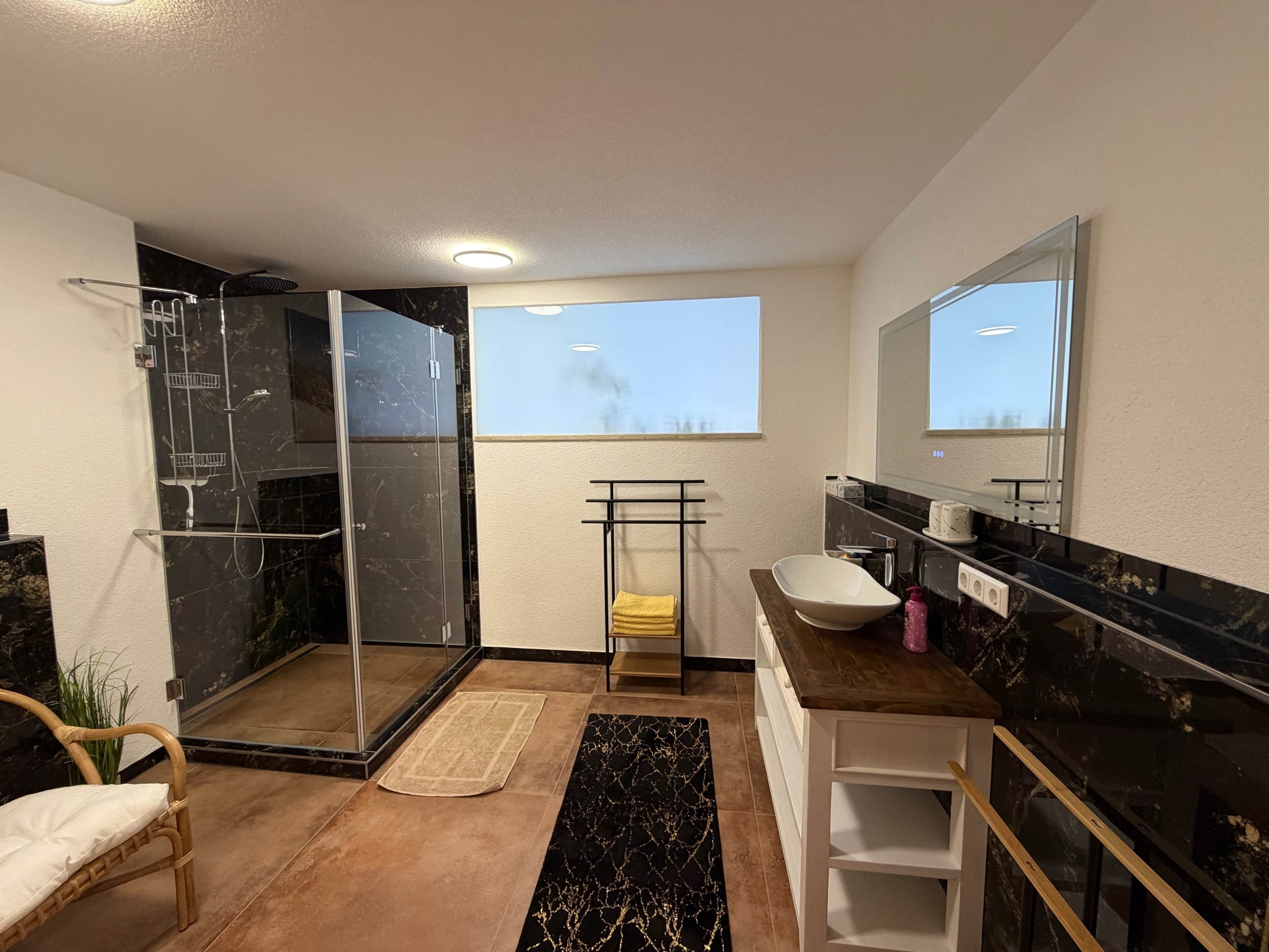 A modern bathroom with a glass shower enclosure, a white sink, a large mirror, and a view of the sky through a window.