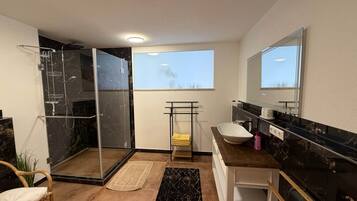 A modern bathroom with a glass shower enclosure, a white sink, a large mirror, and a view of the sky through a window.