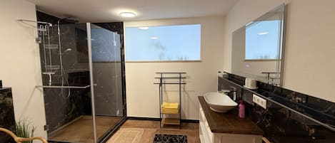 A modern bathroom with a glass shower enclosure, a white sink, a large mirror, and a view of the sky through a window.