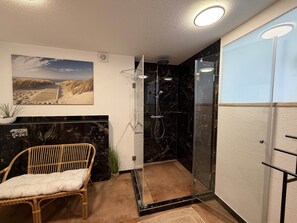 Ein Badezimmer mit einer Duschkabine, einem Korbstuhl, einer Topfpflanze und einem an der Wand befestigten Regal.