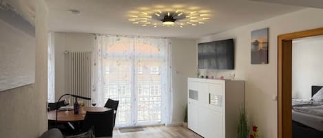 A modern living space with a dining area, a sofa, a TV mounted on the wall, and a bed in the adjacent room.