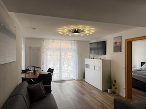 Ein modernes Wohnzimmer mit Essbereich, einer Couch, einem an der Wand montierten Fernseher und einem Bett im angrenzenden Raum.
