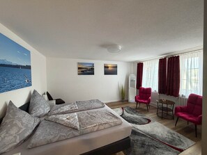 Ein Hotelzimmer mit einem Bett, zwei Gemälden, einem fenster mit Vorhängen, einem Sessel, einem kleinen Tisch und einer Pflanze.