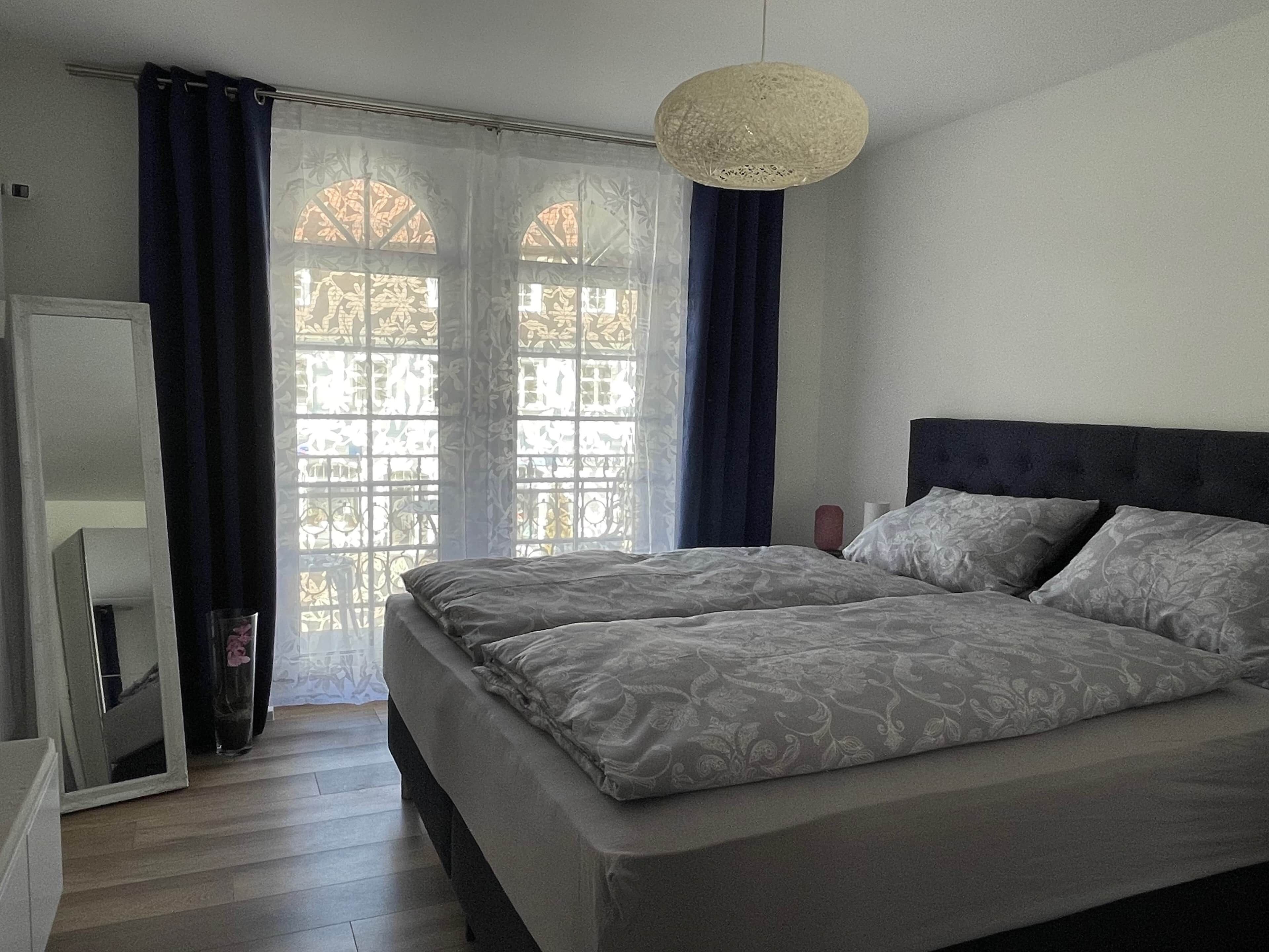 A bedroom with a bed, curtains, a mirror, and a hanging light fixture.