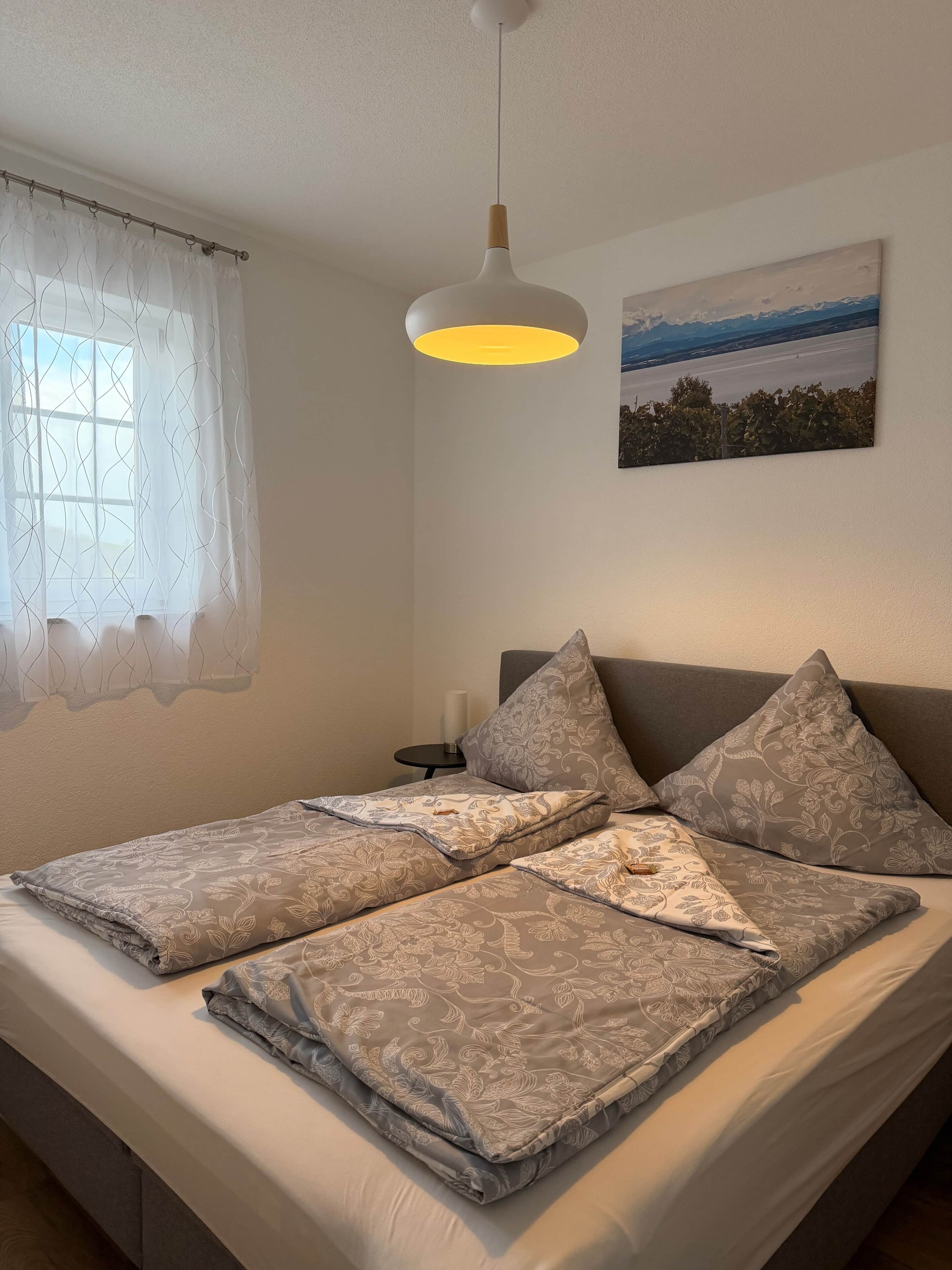 Ein ordentlich bezogenes Bett mit gemusterten Textilien, ein Nachttisch mit Kerze und ein an der Wand hängendes Bild.