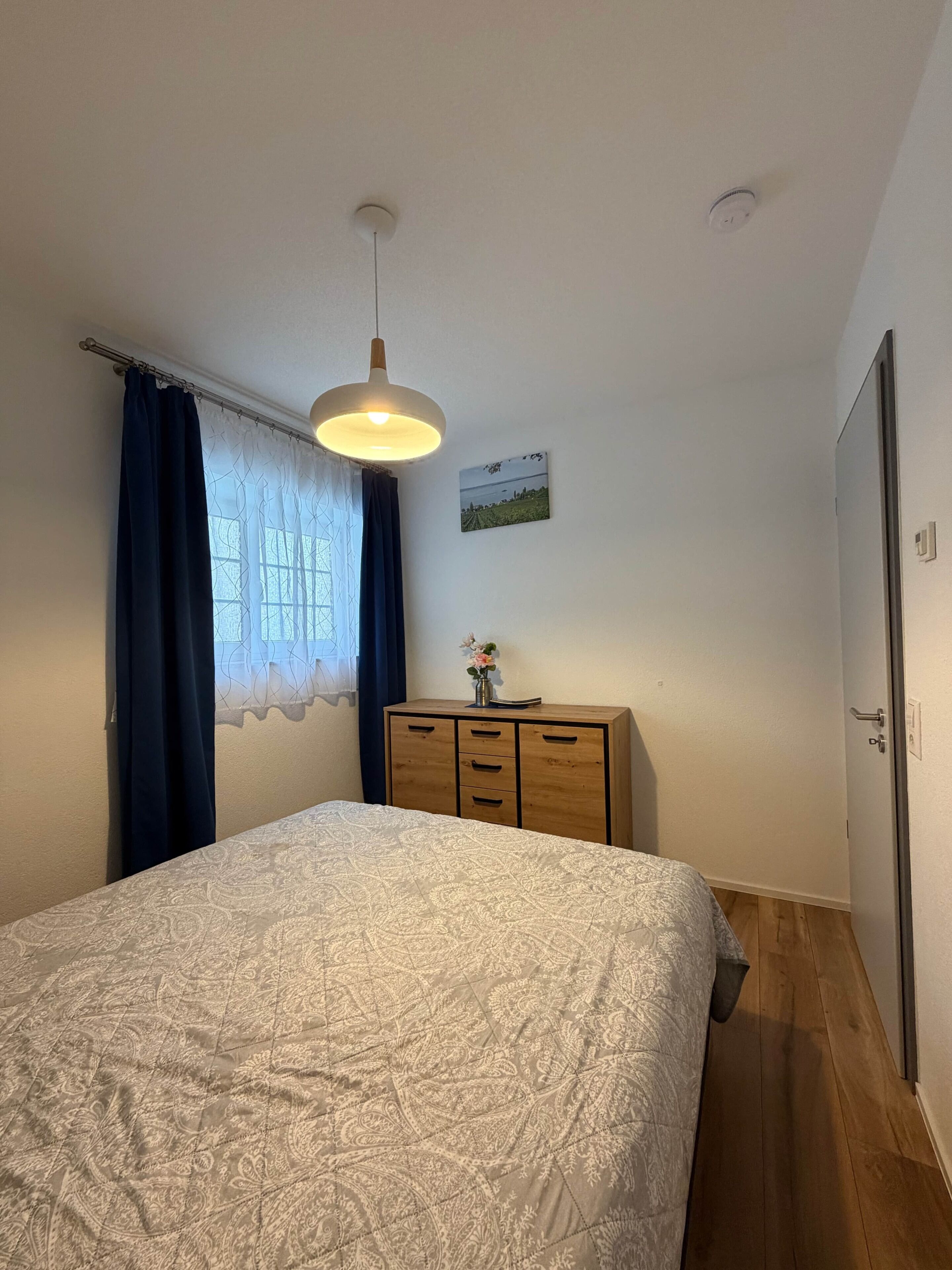 Ein Schlafzimmer mit einem Bett, einer Kommode, einer Hängelampe, einem Fenster mit Vorhängen und einem Wandbild.