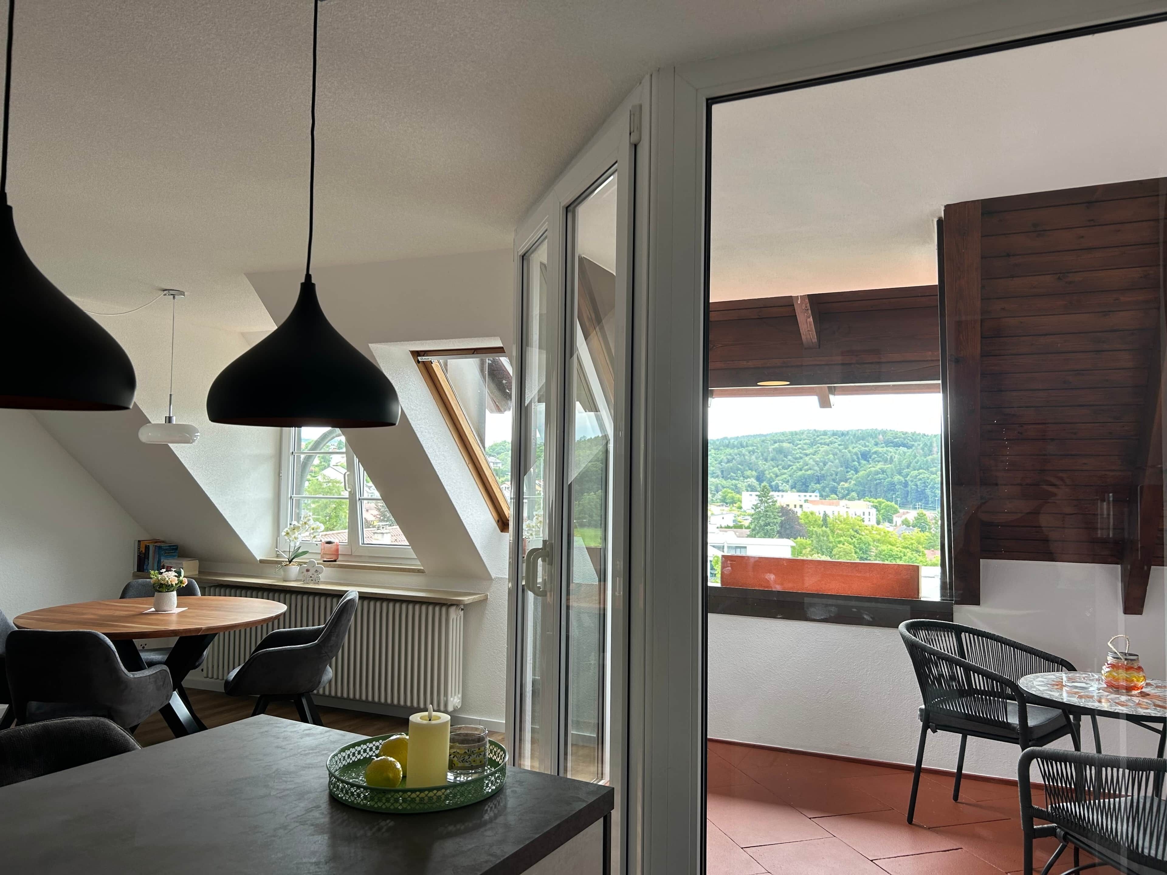A modern dining area with a table, chairs, and a view of a green landscape through large windows.
