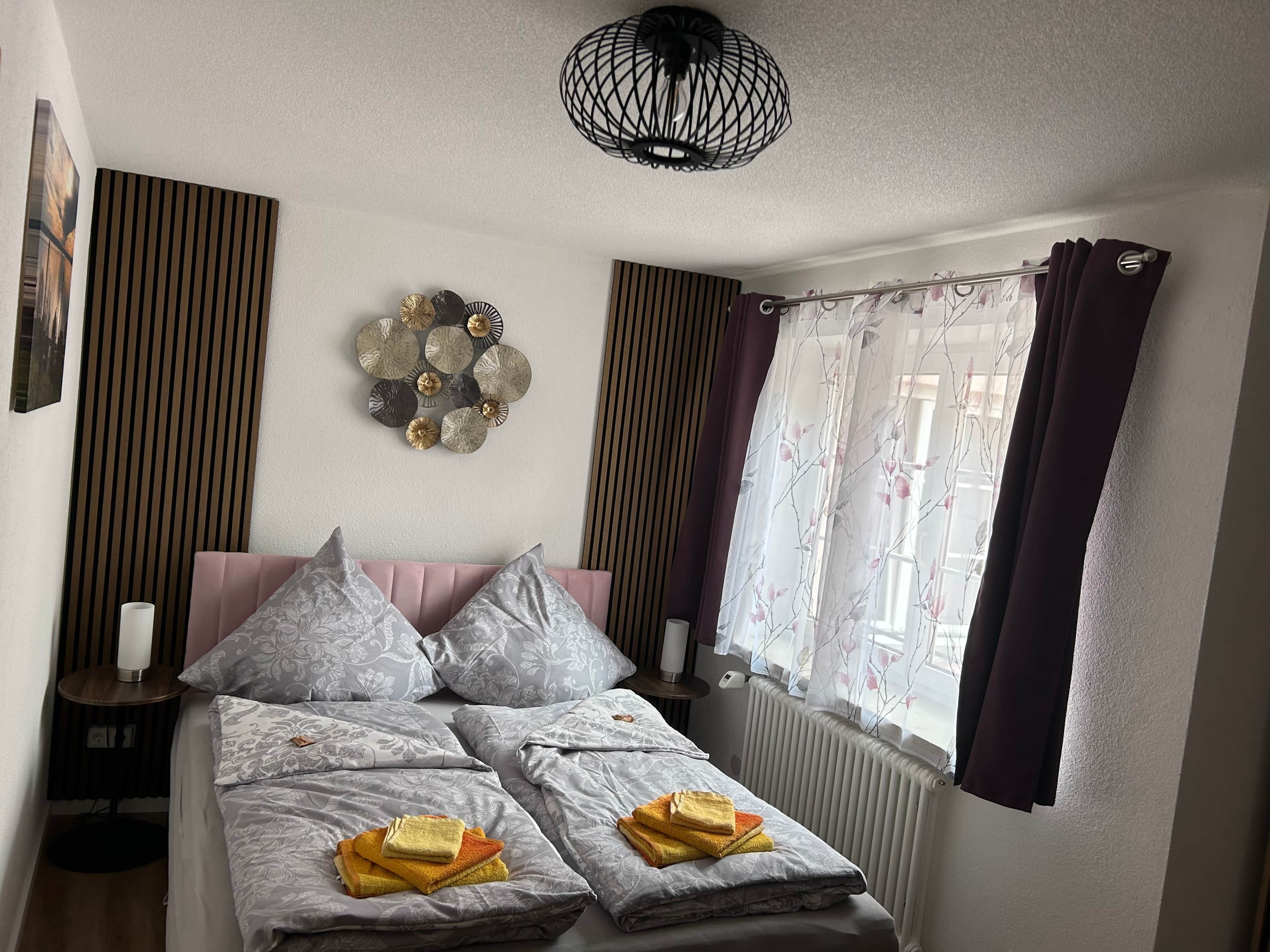 A neatly made bed with pillows and folded towels, a wall-mounted decorative item, a bedside table with a lamp, and a window with curtains.