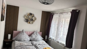 Una cama bien hecha con almohadas y toallas dobladas, un adorno en la pared, una mesita de noche con lámpara y una ventana con cortinas.