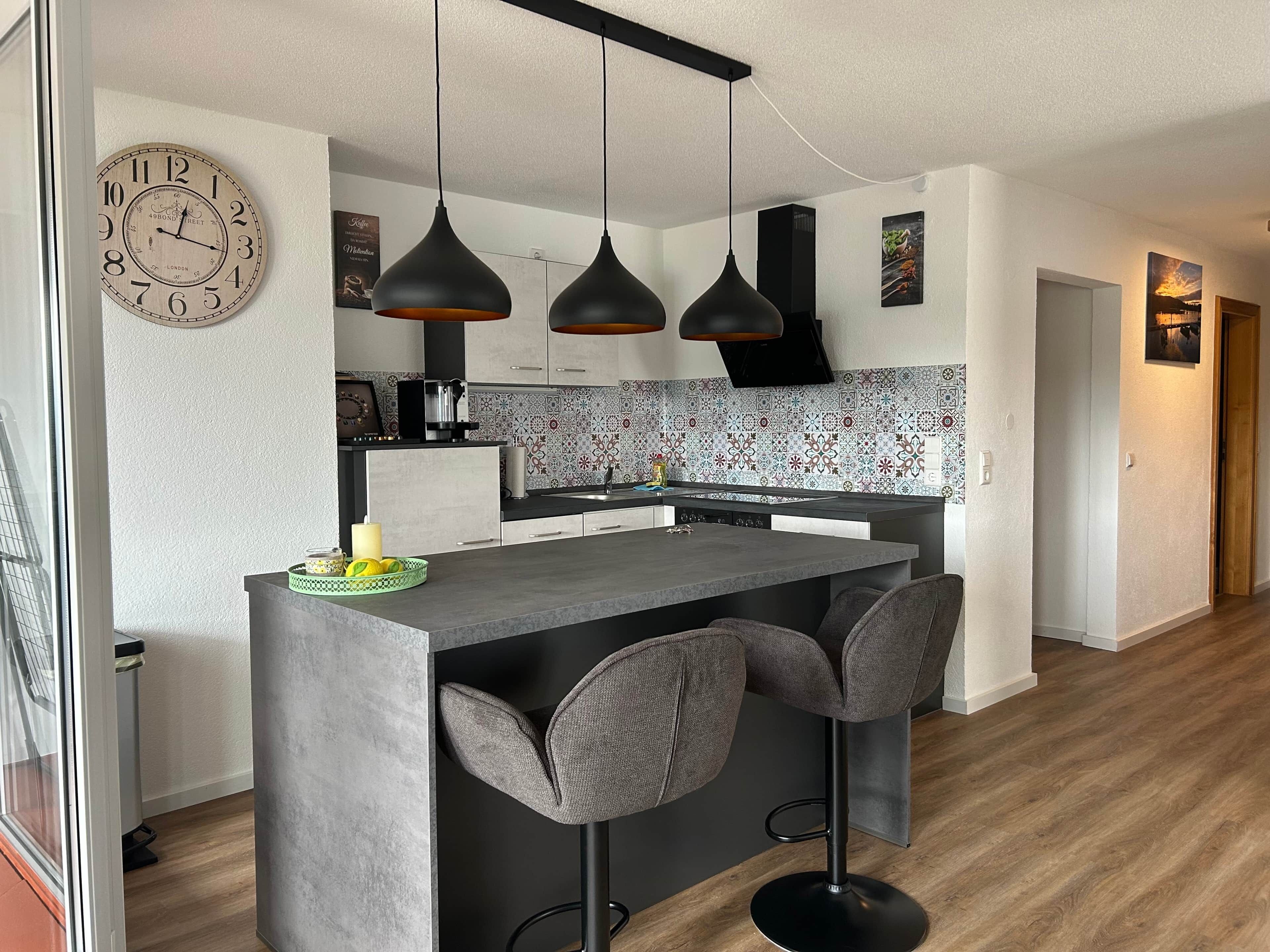 Une cuisine moderne avec un îlot central, des luminaires suspendus et une horloge au mur.