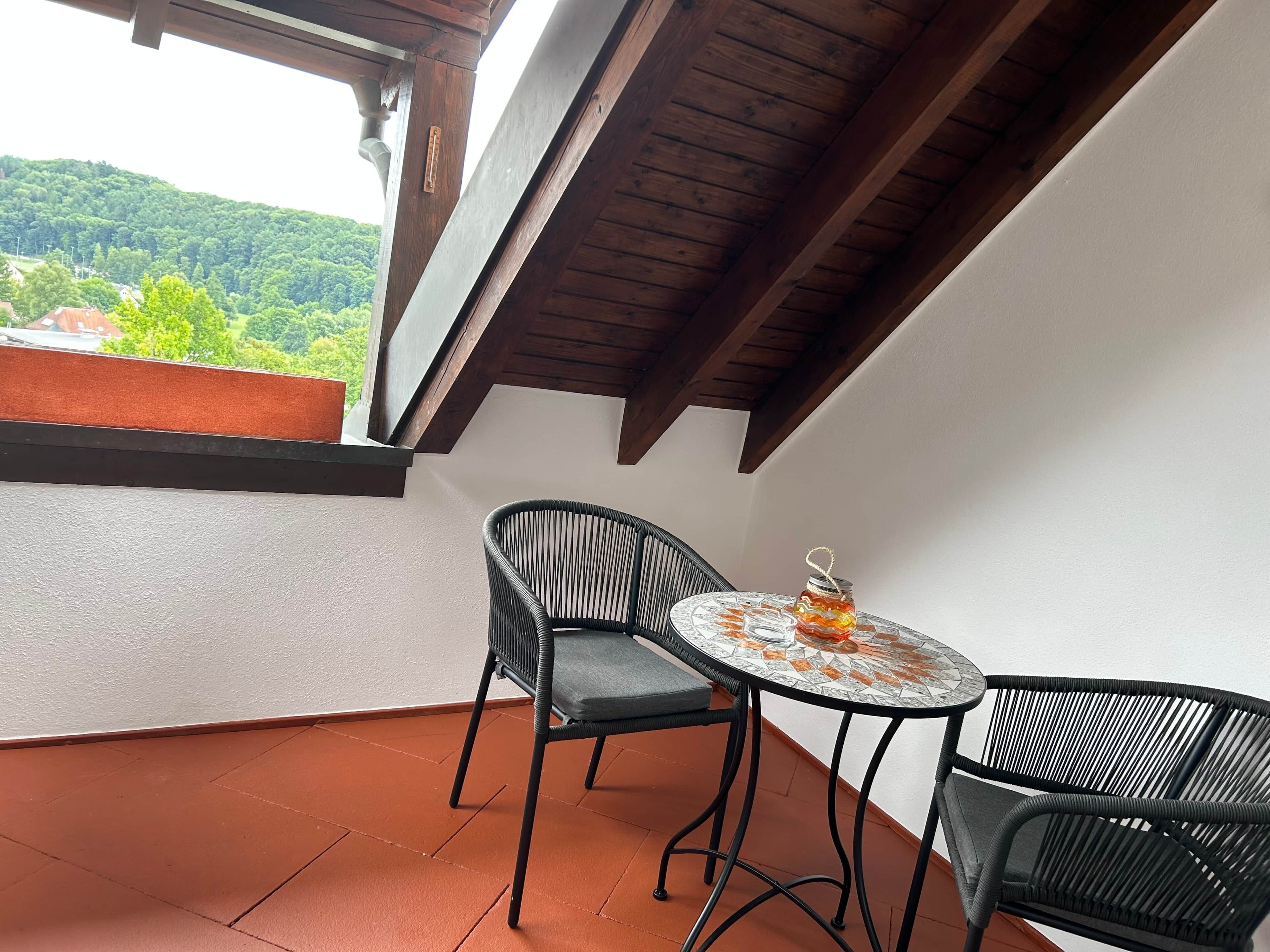A balcony with a table and two chairs, a view of greenery, and a wooden ceiling.