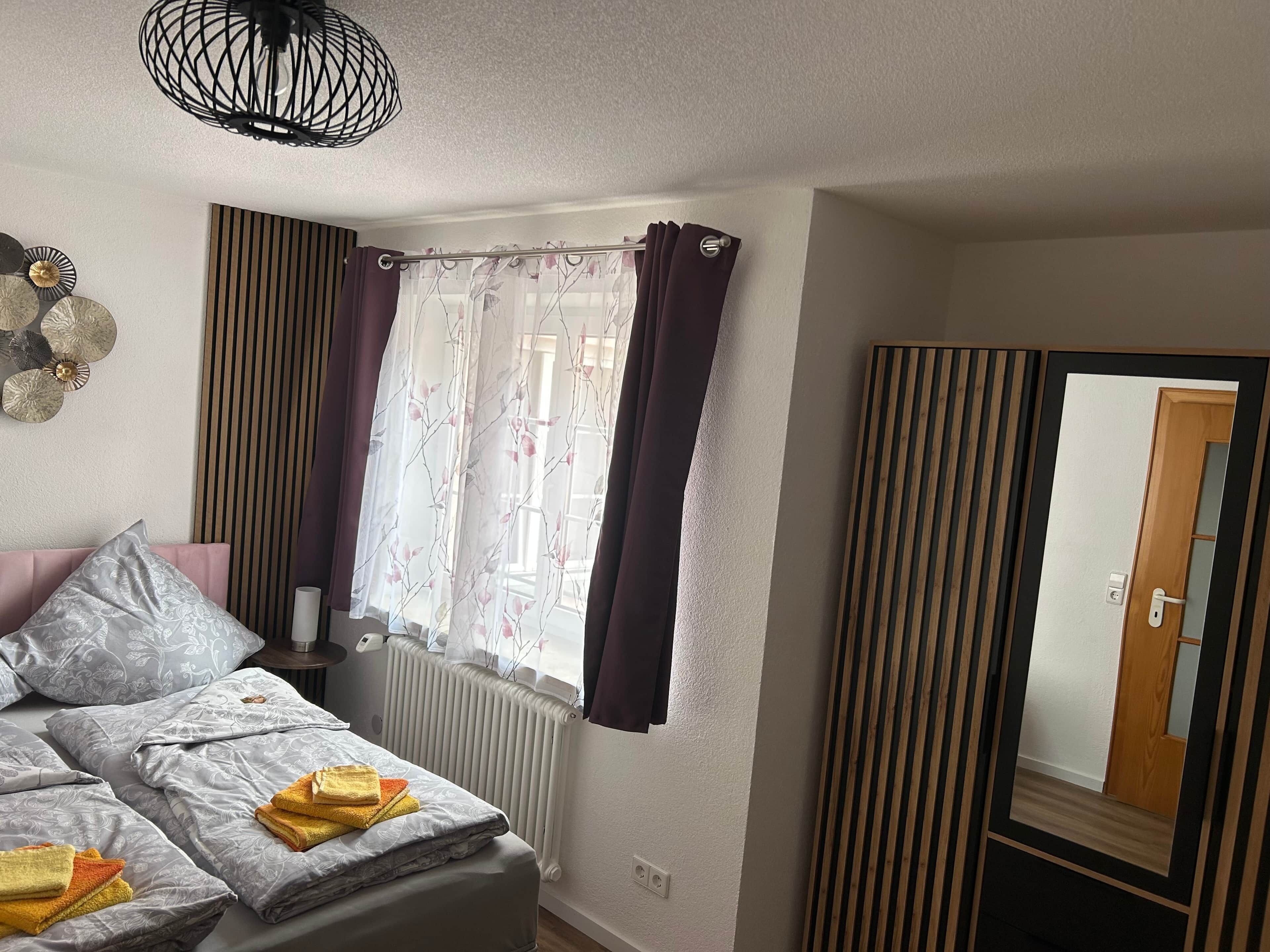 Ein Schlafzimmer mit einem Bett, einem Heizkörper, einem Fenster mit Vorhängen, einem Spiegel und einer Deckenleuchte.