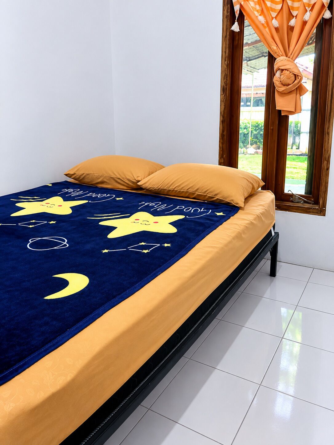 Una cama con colcha azul y amarilla con estrellas y lunas, una ventana con cortinas y un piso de baldosas.