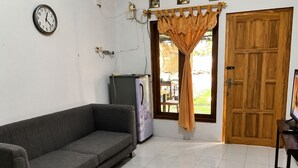 Ein Zimmer mit einer grauen Couch, einer Holztür, einem Fenster mit Vorhängen, einer Wanduhr und einem Kühlschrank.