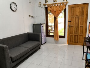Ein Zimmer mit einer grauen Couch, einer Holztür, einem Fenster mit Vorhängen, einer Wanduhr und einem Kühlschrank.