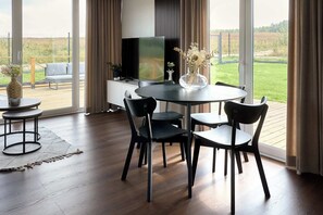 Una sala de estar moderna con una mesa de comedor redonda, sillas negras, un televisor de pantalla plana y vistas a un área verde exterior a través de grandes ventanales.