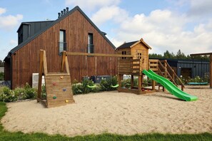 A wooden play structure with a slide and climbing wall, set against a backdrop of a large barn and a smaller wooden building.
