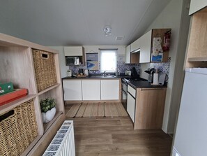 A compact kitchen with white cabinets, a microwave, a sink, and a stove. There are wicker baskets on shelves and a small plant on the counter. - Demelza Seven Bays Holiday Park (Padstow)