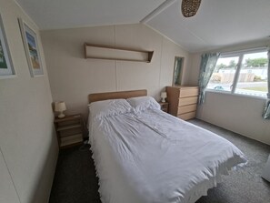 A bedroom with a bed, bedside table, dresser, and a window with curtains. - Demelza Seven Bays Holiday Park (Padstow)