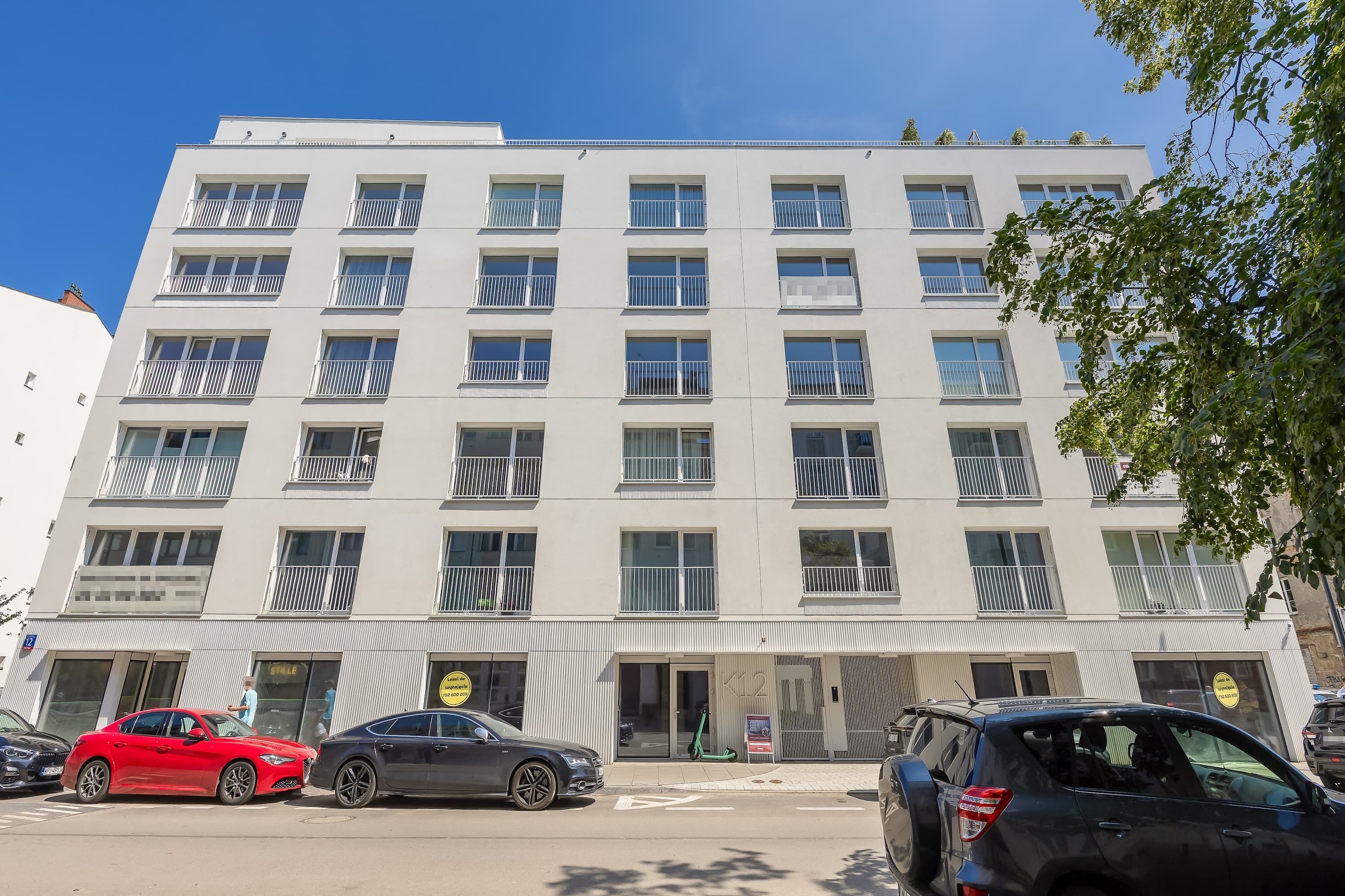 A multi-story building with balconies, a parking area with cars, and a clear sky.