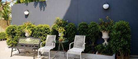 An outdoor seating area with a bench and two chairs, a small table, and potted plants against a dark wall.