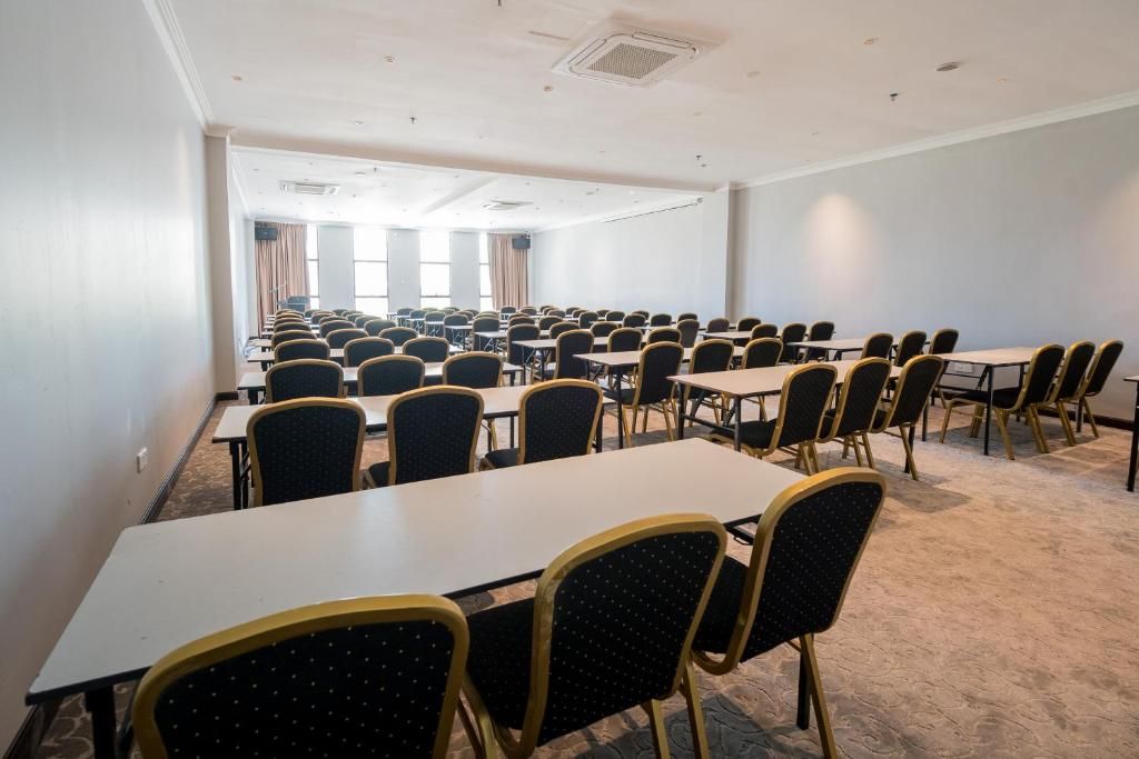 Uma sala de conferências com mesas e cadeiras dispostas em fileiras, amplas janelas com cortinas e piso coberto por carpete.