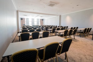 Uma sala de conferências com mesas e cadeiras dispostas em fileiras, amplas janelas com cortinas e piso coberto por carpete.