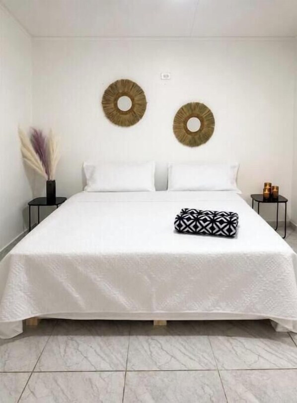 Un lit recouvert d’une couvre-lit blanche, avec deux oreillers et un coussin décoratif, le tout placé devant un mur blanc.