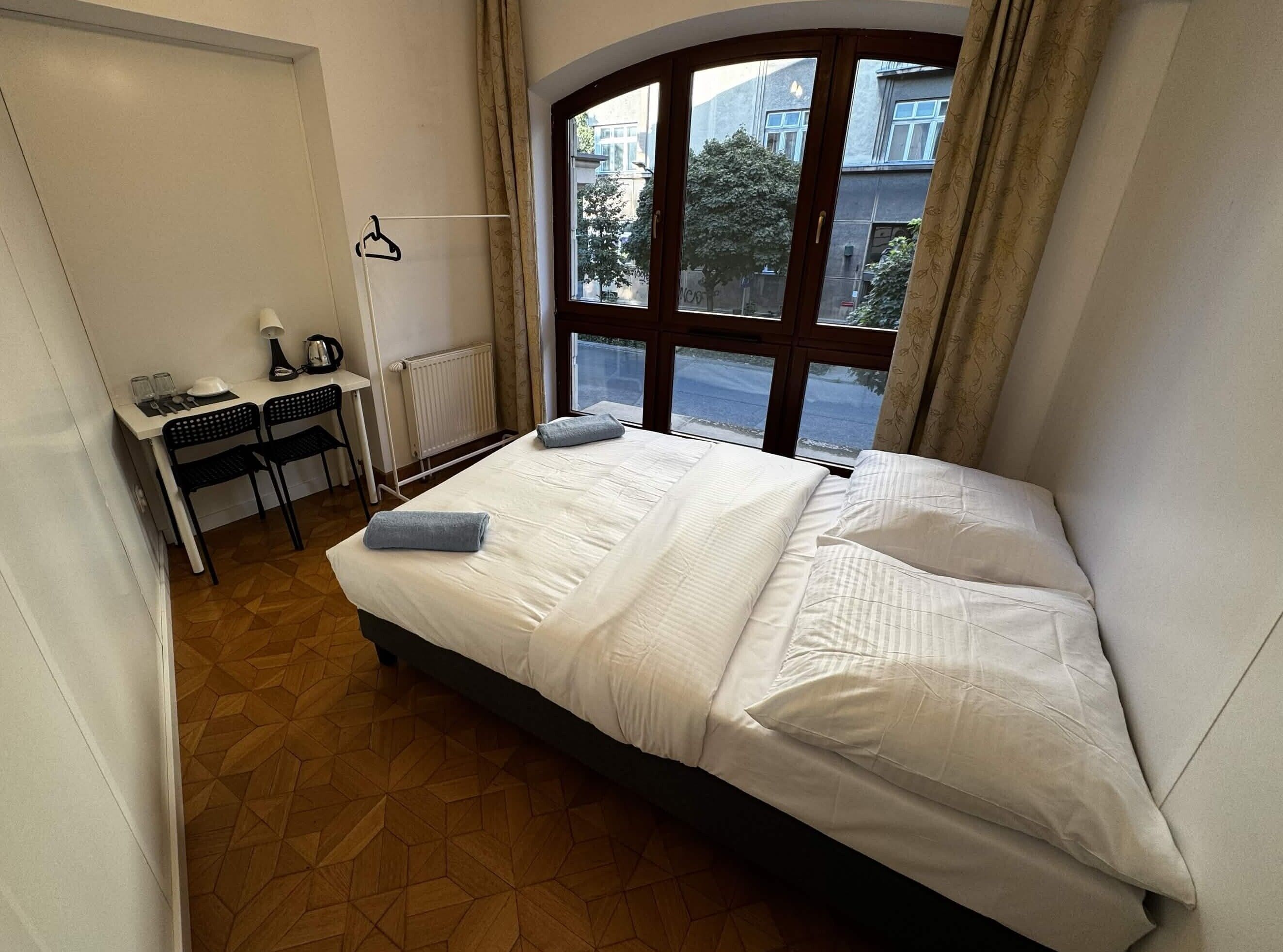 A hotel room with a bed, a desk, a chair, and a window with a view of buildings and trees.