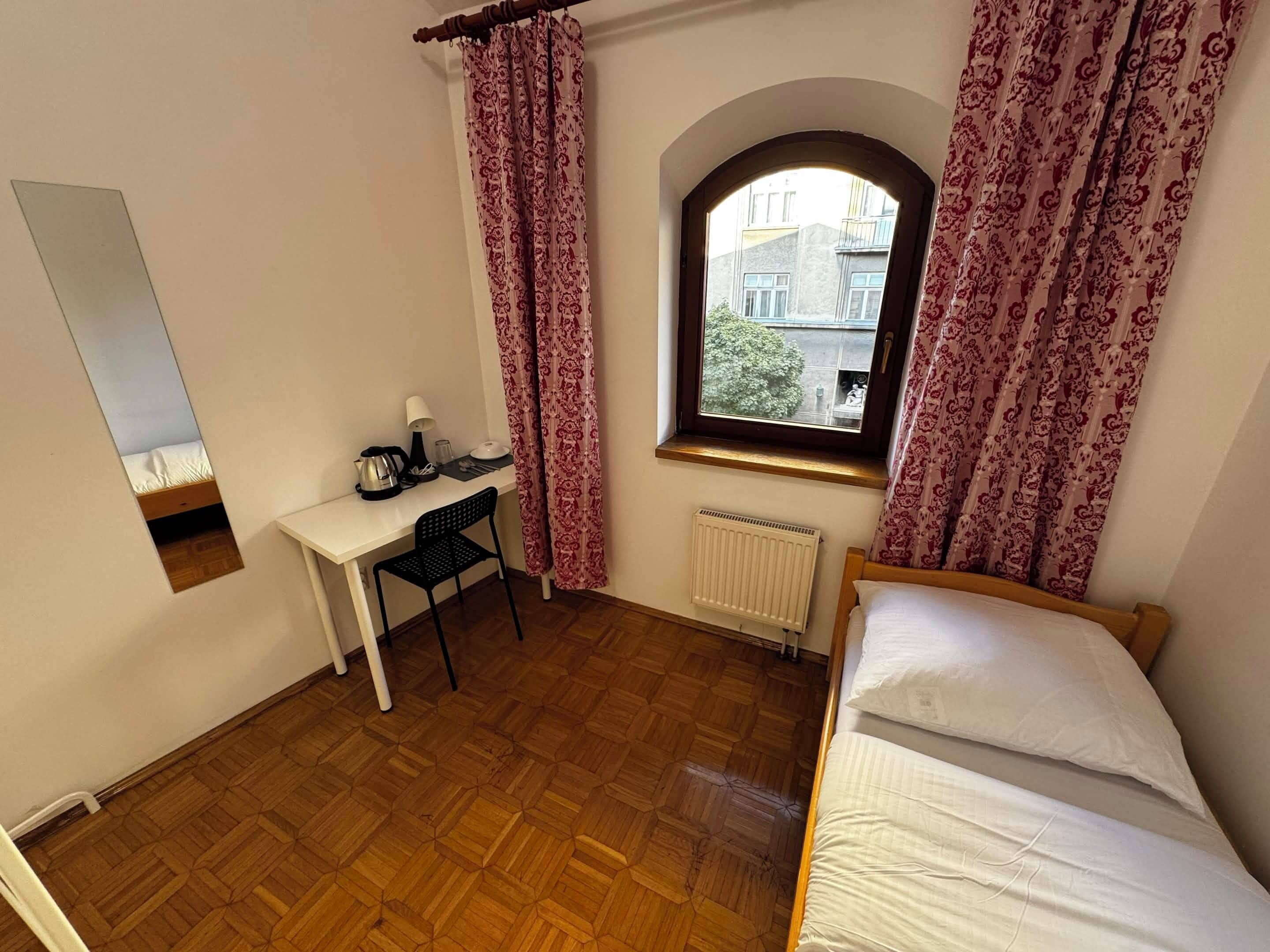 Une petite pièce avec un plancher en bois, un lit, un bureau avec une chaise, un radiateur et une fenêtre avec des rideaux.