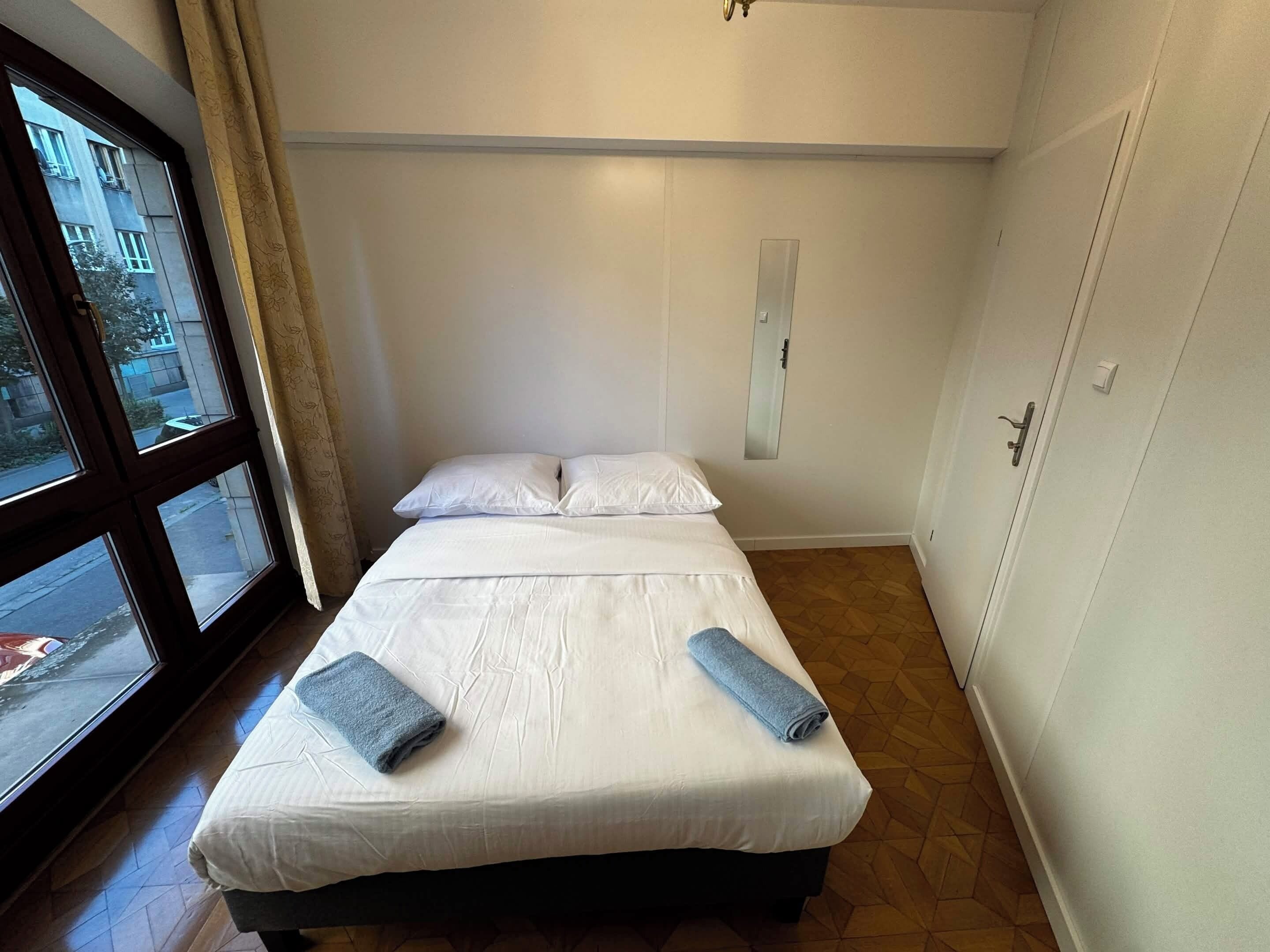 A single bed with two pillows and two rolled towels, a window with curtains, and a door with a handle.