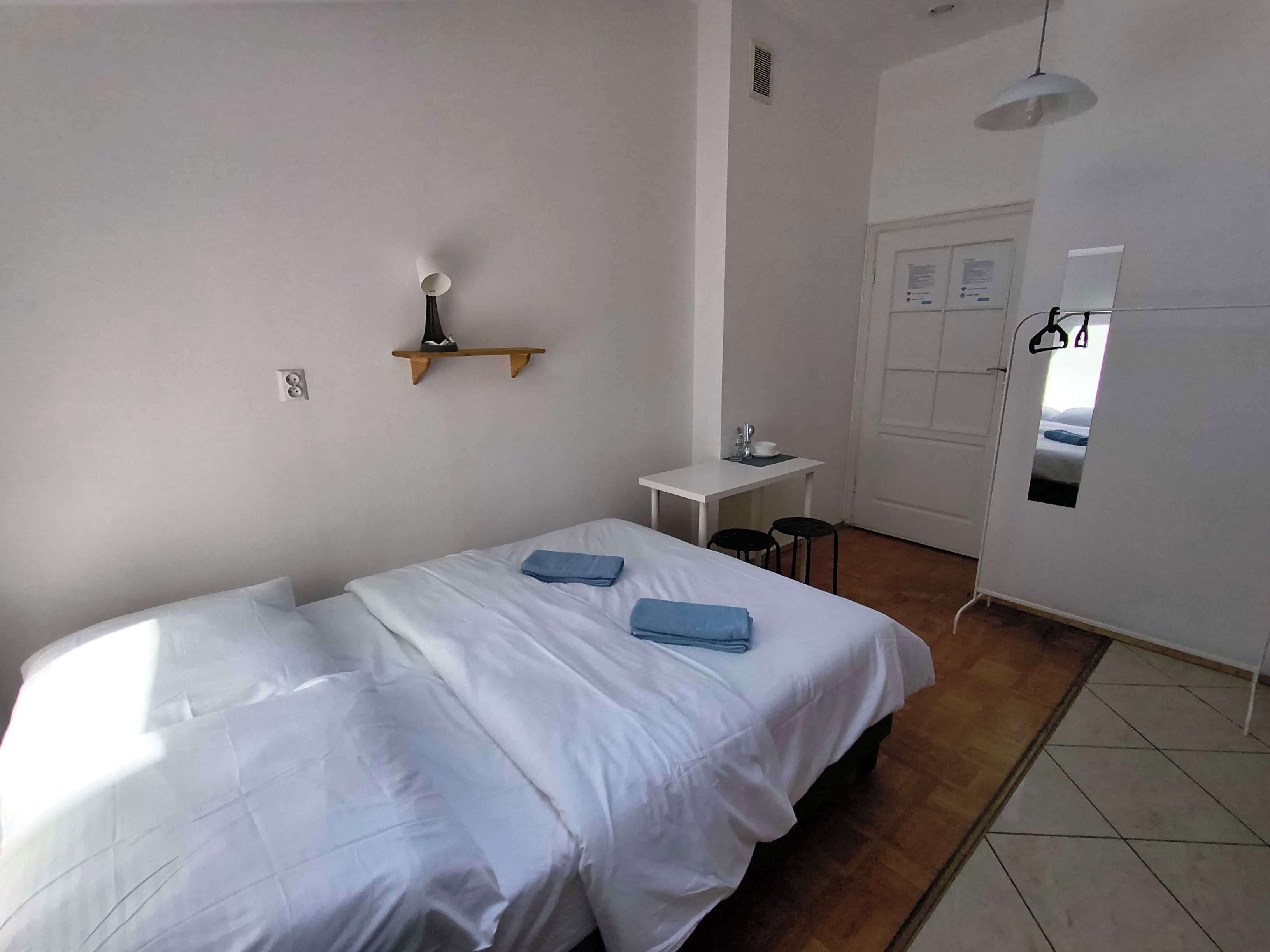 Een hotelkamer met een bed, een klein bureau, een stoel, een plank met een lamp en een deur met een spiegel.