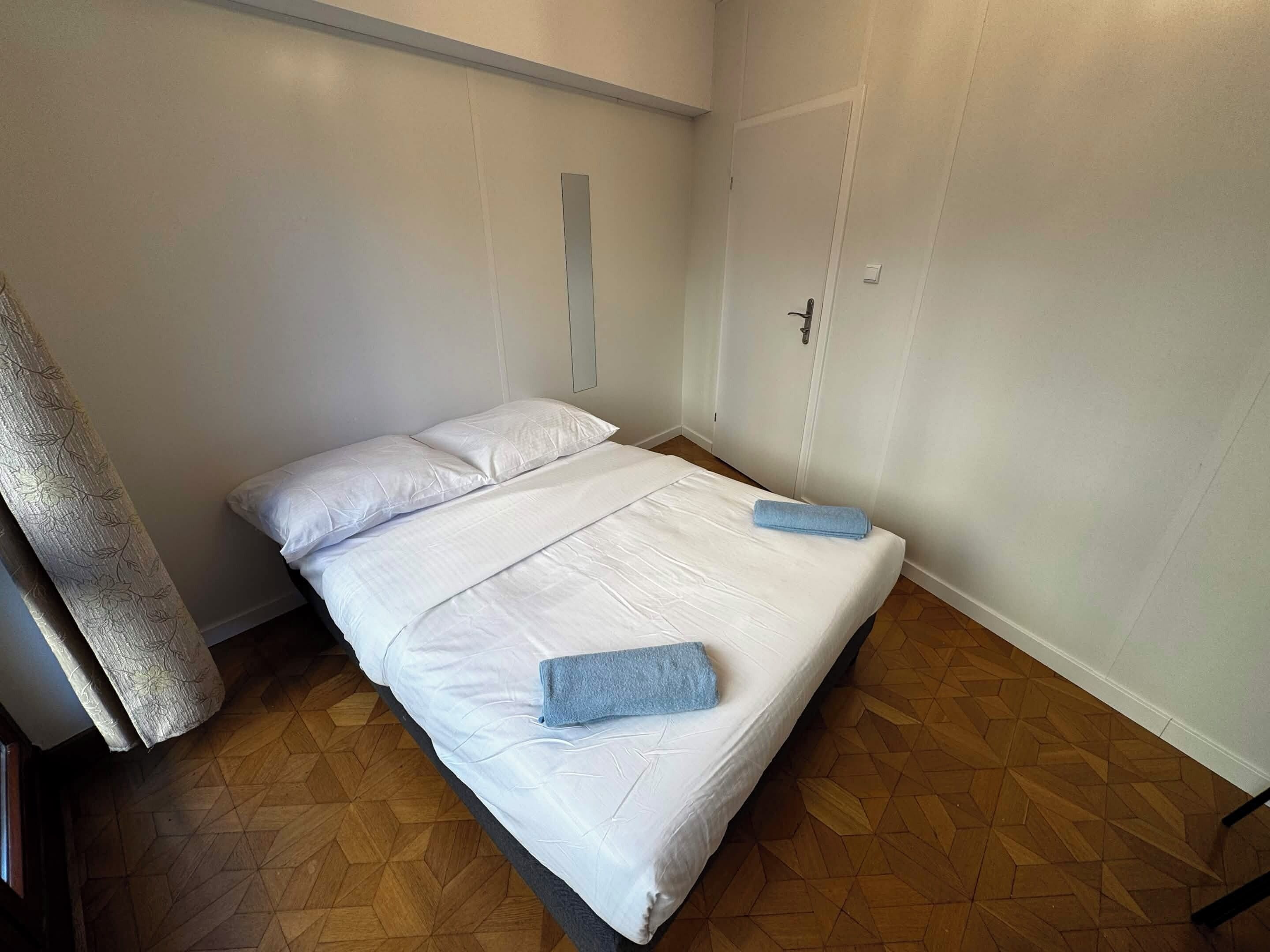 Un lit simple avec du linge de lit blanc et deux serviettes bleues, un plancher en bois et une porte blanche avec une poignée.