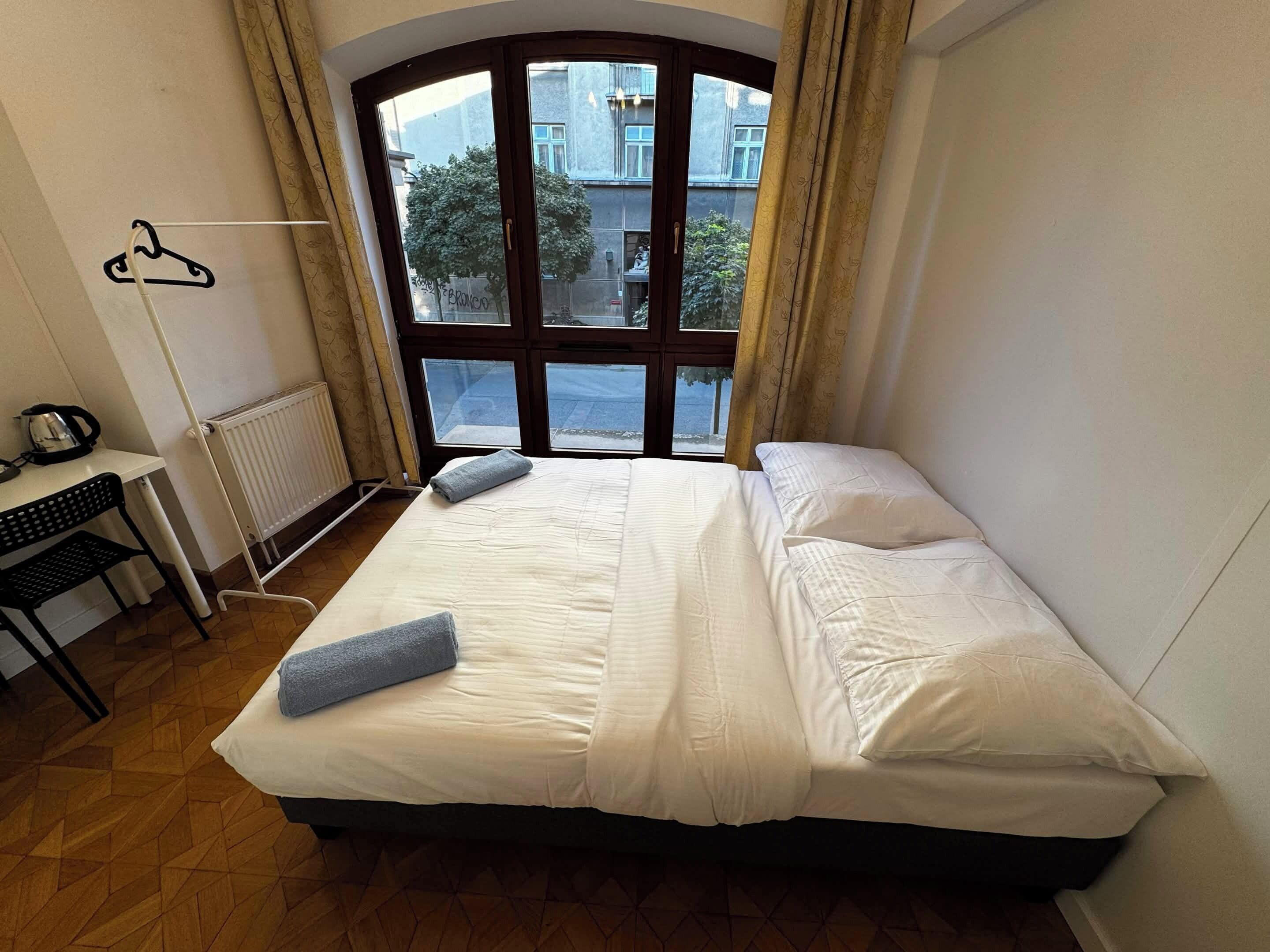 Une chambre à coucher avec un lit, un radiateur, un bureau avec une bouilloire et une fenêtre avec des rideaux.