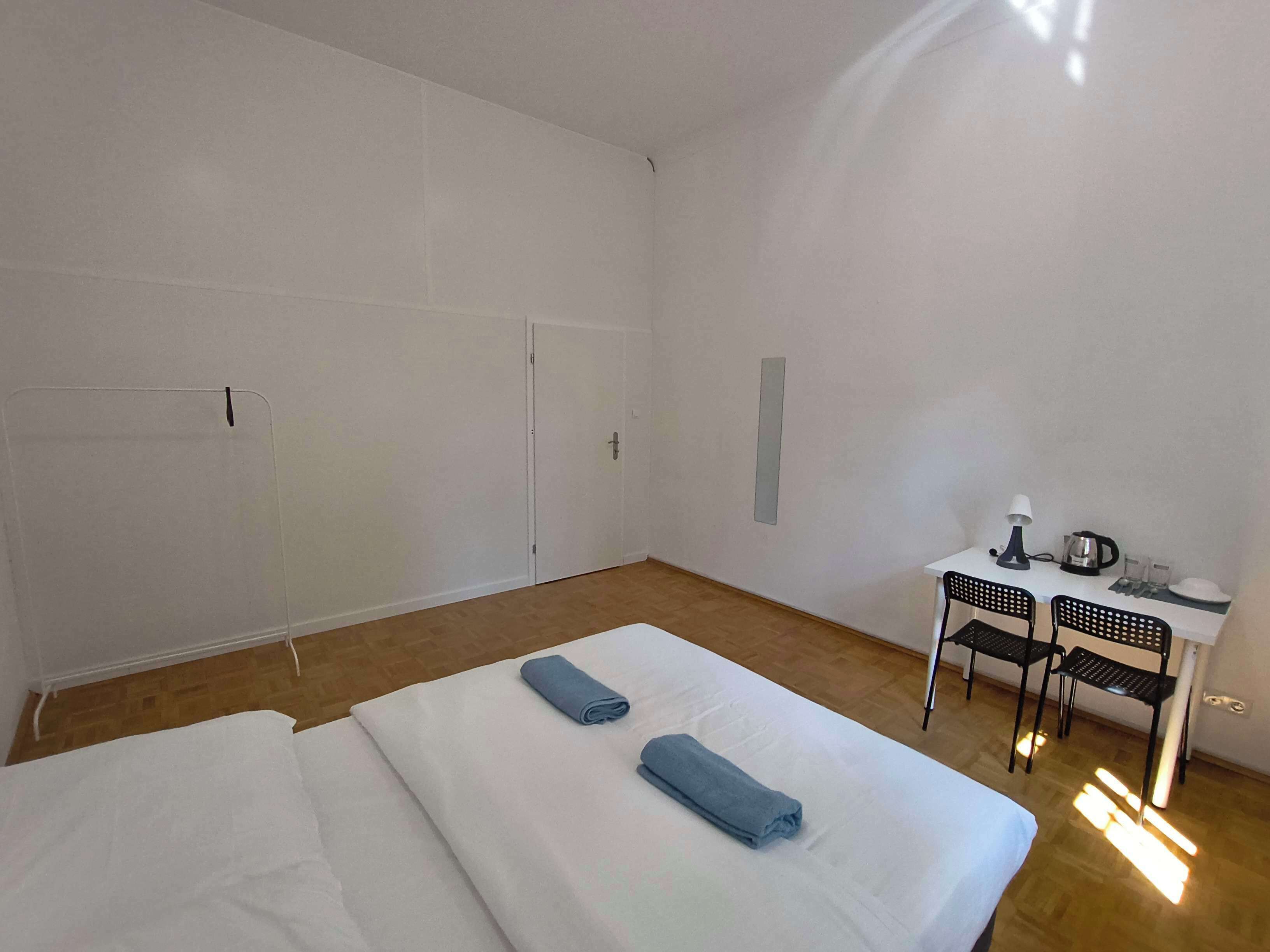 A hotel room with a bed, two rolled towels, a desk with a kettle, and two chairs.