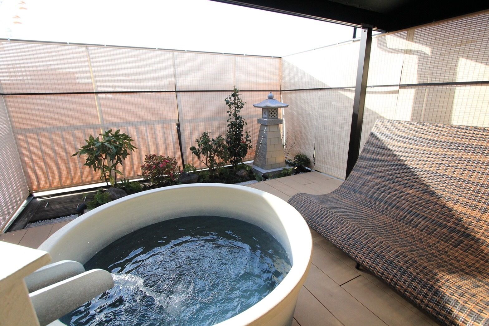 A hot tub with a stone surround, a woven sofa, a small garden with plants and a lantern, and a mesh fence.