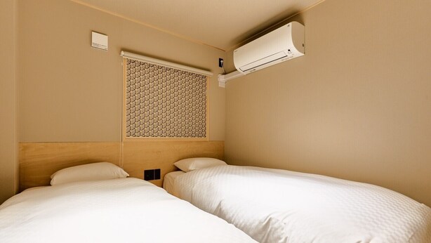 A hotel room with two beds, a window with a patterned blind, and an air conditioning unit mounted on the wall. - Imakusa-so, Osaka, Tempozan - Tall Lantern - (大阪府大阪市港区築港3丁目)