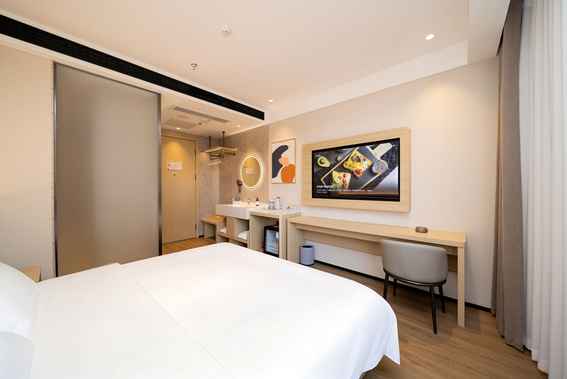 A hotel room with a bed, a desk, a chair, a mirror, and a wall-mounted TV.