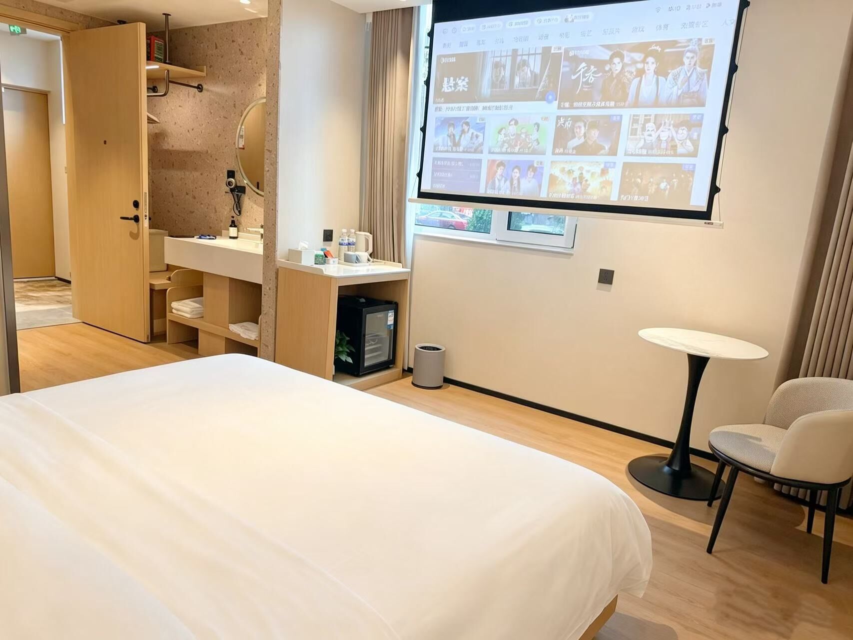 Ein Hotelzimmer mit Bett, Schreibtisch, Stuhl, einer Projektionsleinwand, die Bilder zeigt, und einem kleinen Tisch.