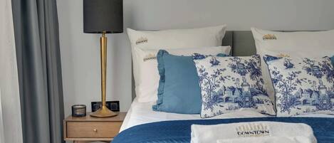 Una habitación de hotel con cama, mesita de noche, lámpara y ventana con cortinas.