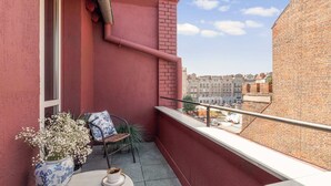 Ein Balkon mit roter Wand, einem Fenster, einem Stuhl, einem Tisch mit Tasse und einem Blumenvasen.