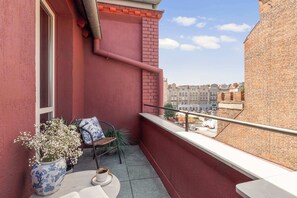 A balcony with a red wall, a window, a chair, a table with a cup, and a vase with flowers. - Lawendowa Room Nº5 | Shared Kitchen | Balcony (Gdańsk)