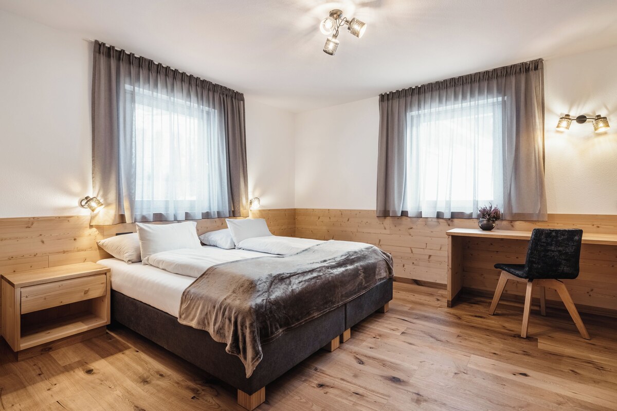 A bedroom with a wooden headboard, a bed with white linens, a bedside table, a desk with a chair, and two windows with curtains.