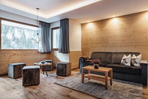 A modern living room with a wooden floor, a dark sofa, a wooden coffee table, and a large window with curtains. - Hotel Alcialc (La Valle)
