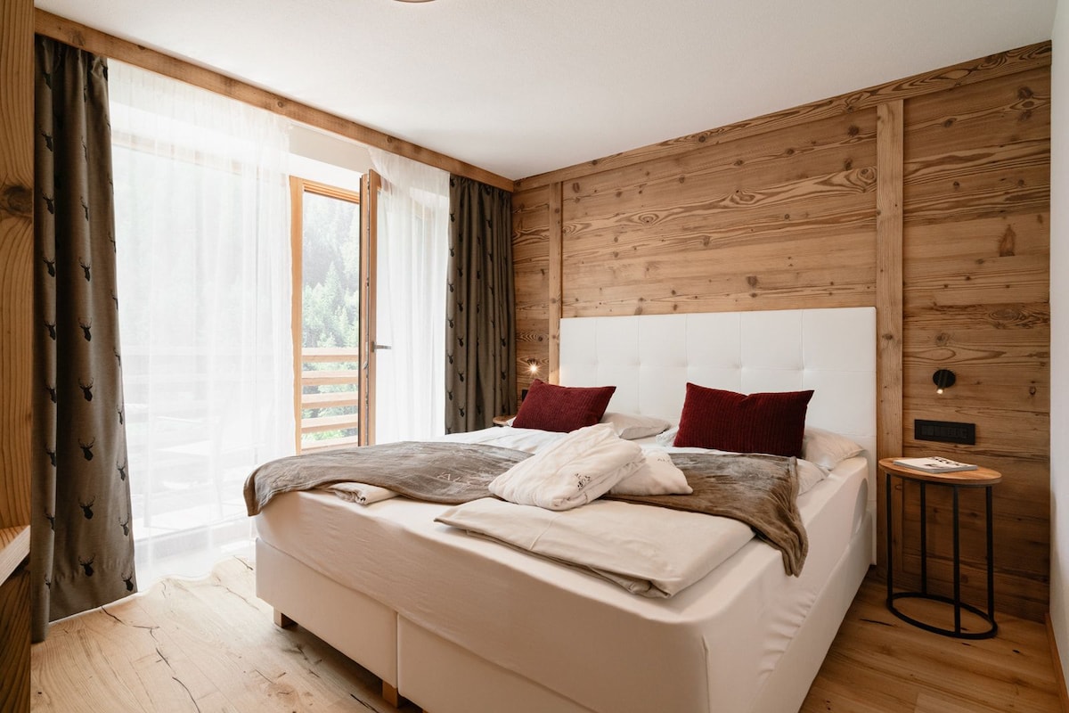A bedroom with a large bed, wooden panel walls, a window with curtains, and a bedside table with a lamp.
